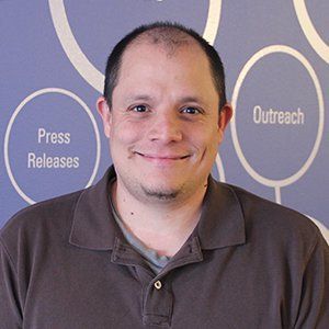 The image shows a man with short hair, smiling, wearing a brown polo shirt, standing in front of a background with circles containing text such as "Press Releases" and "Outreach."